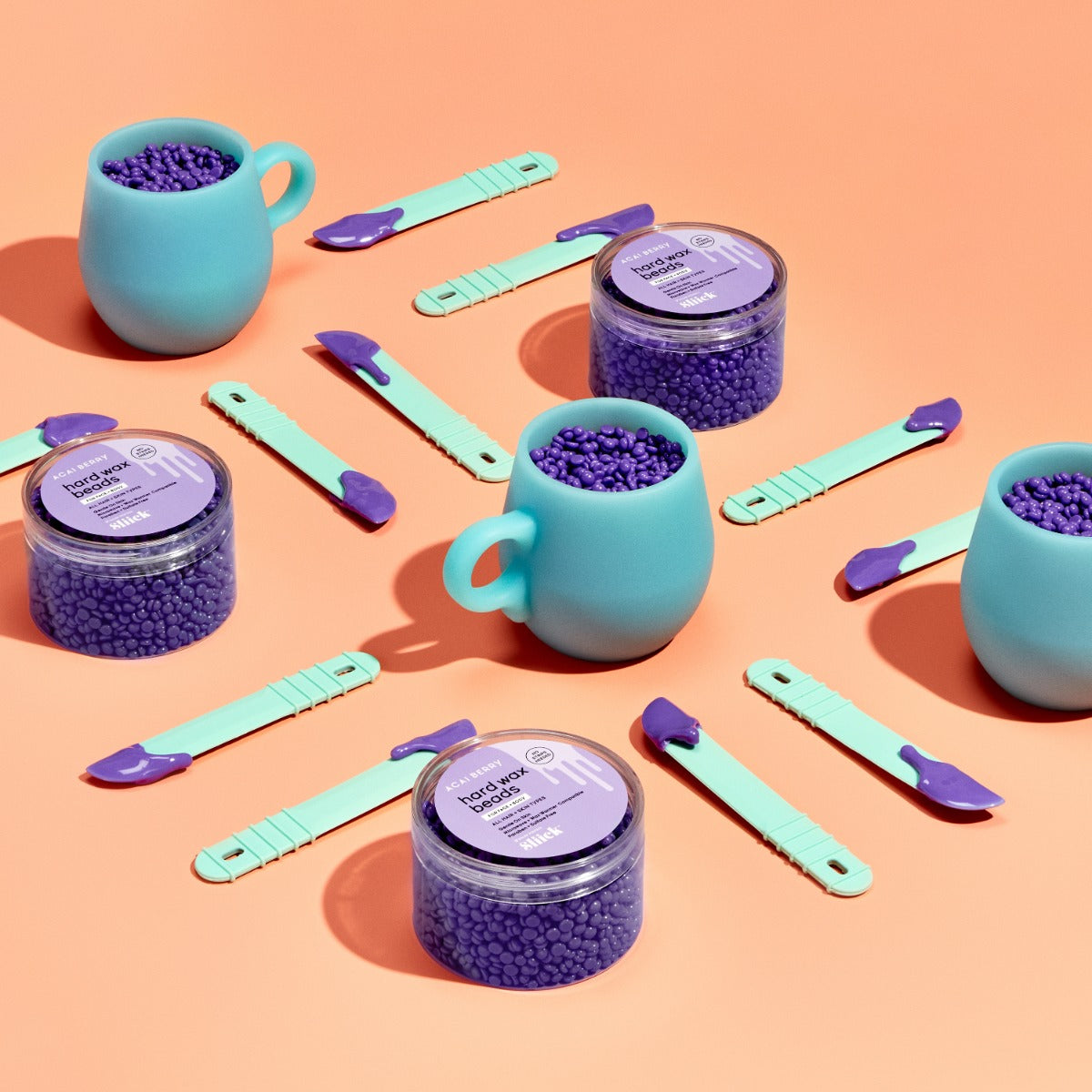 Sliick Hard Wax Flat Lay – Multiple pastel blue melting cups, purple wax beads, open jars, & mint spatulas arranged on a warm peach background.